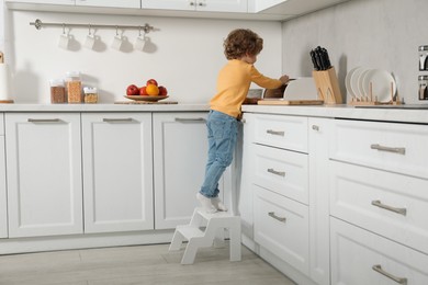 Photo of Little boy standing on step stool and reaching towards bread box in kitchen