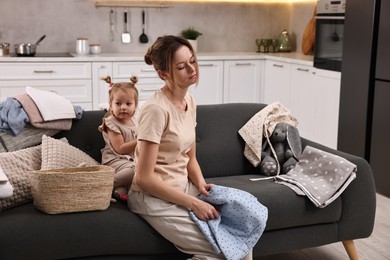 Photo of Tired housewife with her little daughter among messy laundry on sofa at home