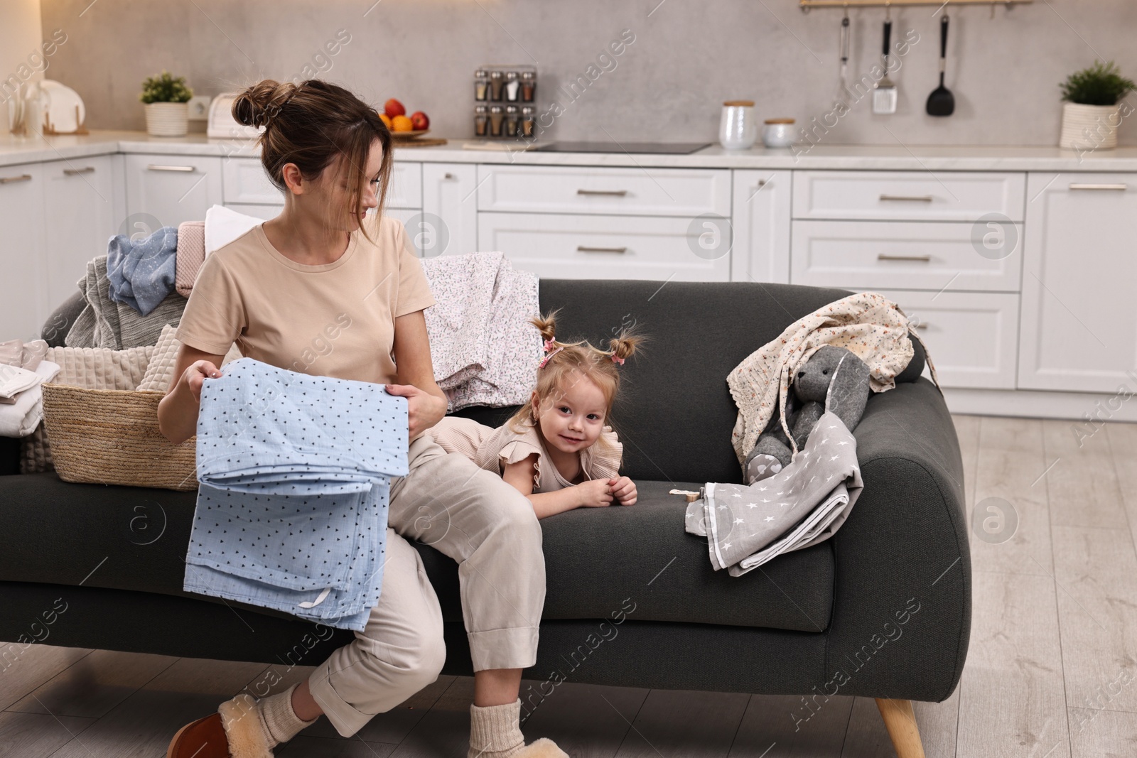 Photo of Housewife with her little daughter among messy laundry on sofa at home