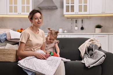 Photo of Housewife with her little daughter among messy laundry on sofa at home