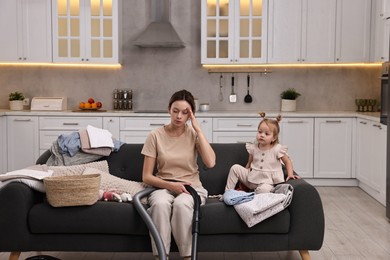 Photo of Tired housewife with her little daughter among messy laundry on sofa at home