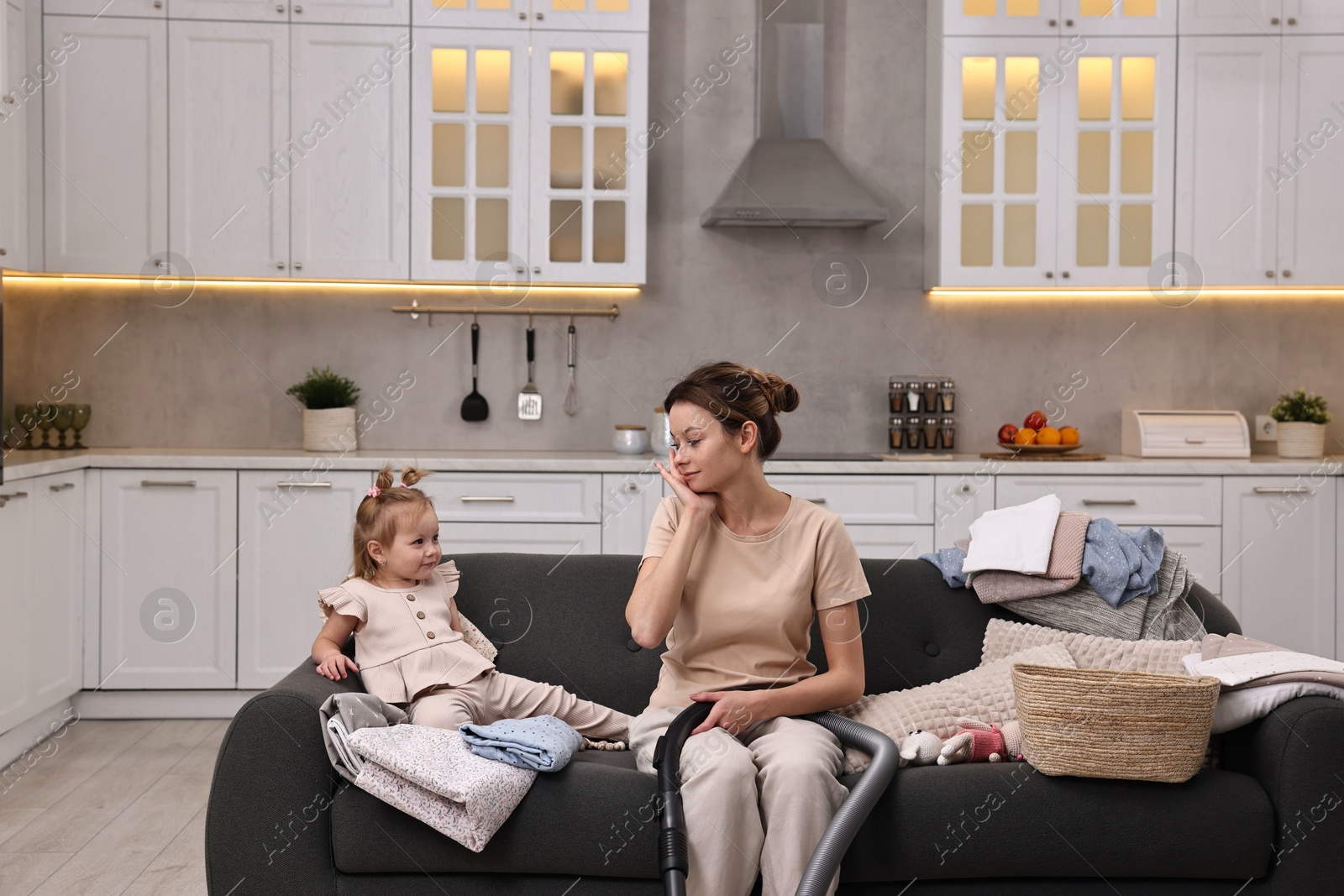 Photo of Tired housewife with her little daughter among messy laundry on sofa at home