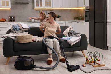 Photo of Housewife with her little daughter looking at something among messy laundry on sofa indoors