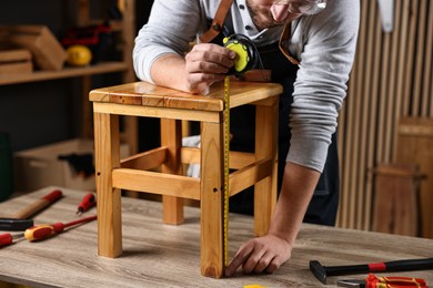 Photo of Repairman measuring wooden stool at table in workshop, closeup