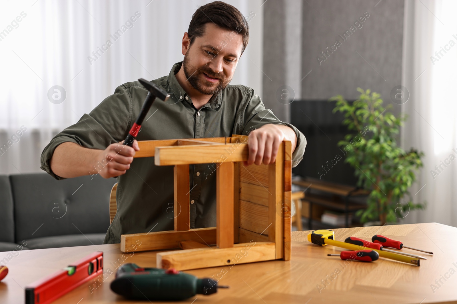 Photo of Man repairing wooden stool with nail and hammer at home