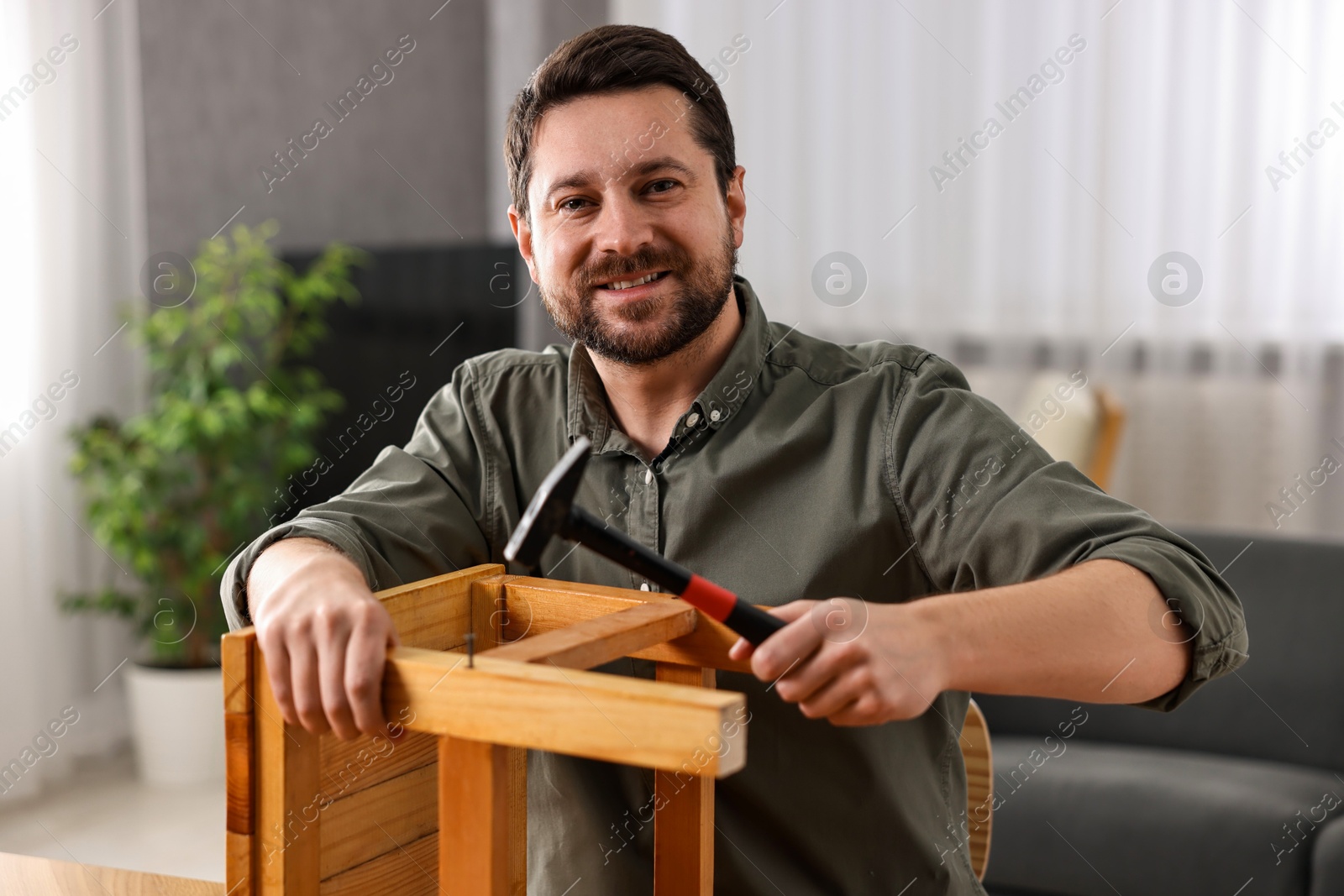 Photo of Man repairing wooden stool with nail and hammer at home