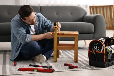 Photo of Man using tape measure while repairing wooden stool at home