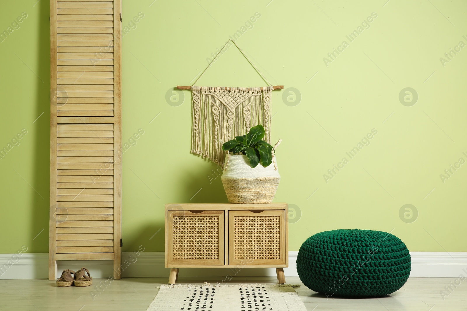 Photo of Stylish room interior with comfortable knitted pouf, wooden furniture and plant near light green wall