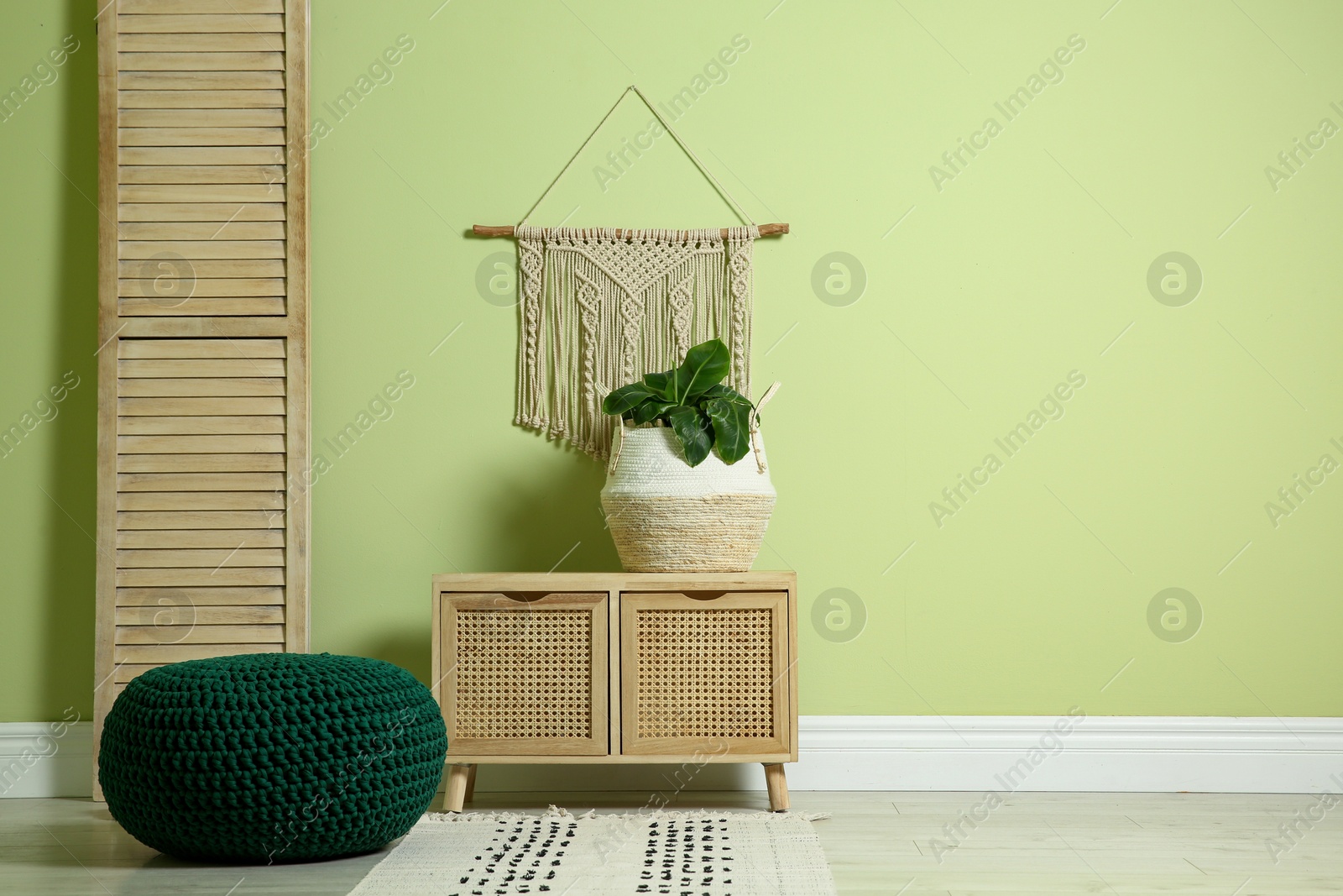 Photo of Stylish room interior with comfortable knitted pouf, wooden furniture and plant near light green wall