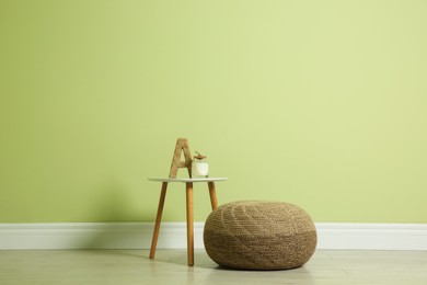 Photo of Comfortable knitted pouf and table with decor elements near light green wall indoors, space for text. Interior design