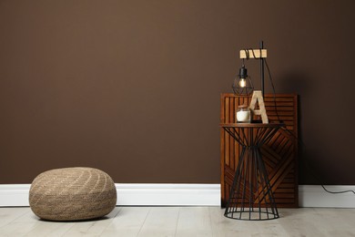 Photo of Comfortable knitted pouf and table with decor elements near brown wall indoors, space for text. Interior design