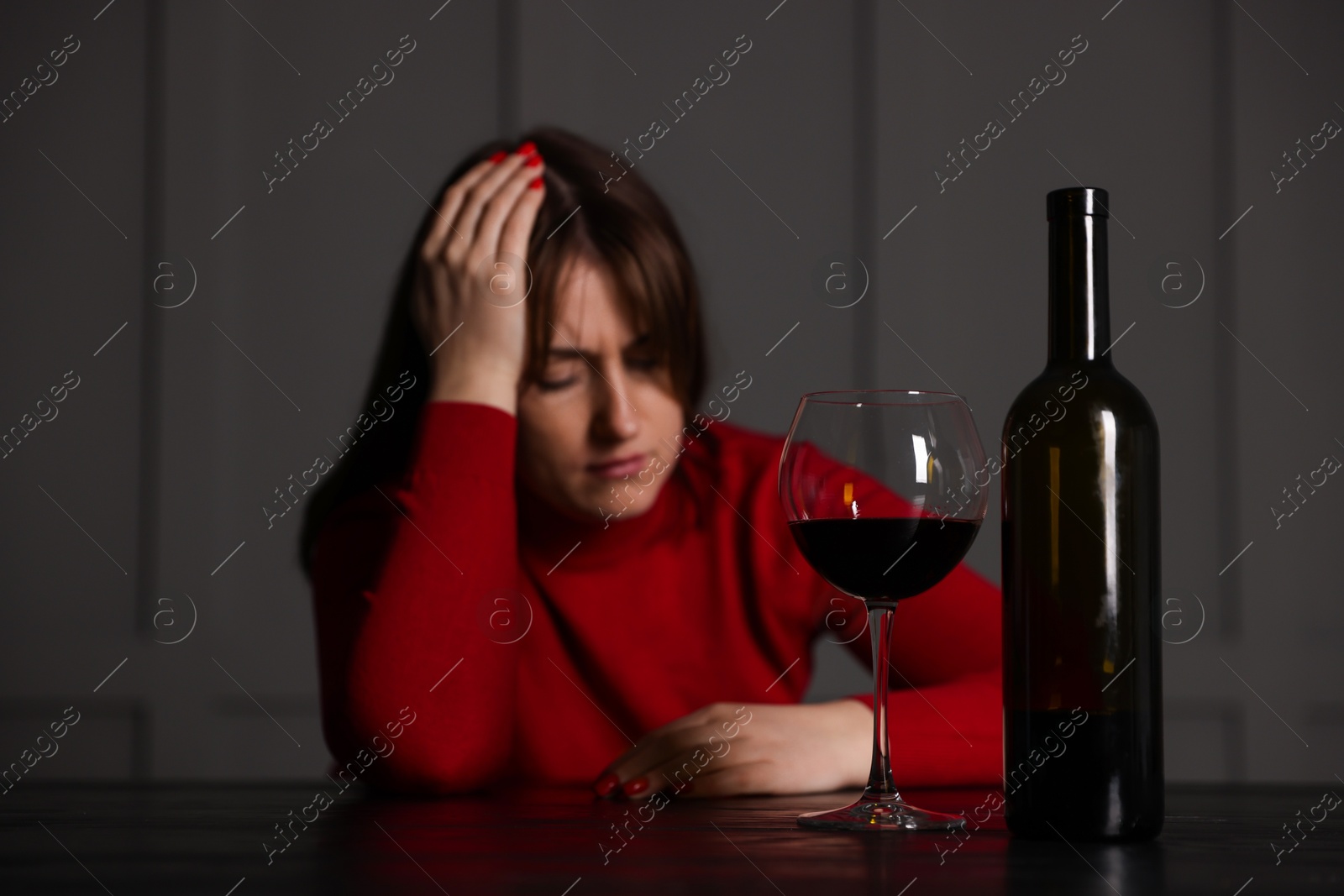 Photo of Alcohol addiction. Woman at wooden table indoors, focus on glass of red wine and bottle