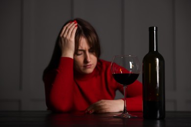 Photo of Alcohol addiction. Woman at wooden table indoors, focus on glass of red wine and bottle