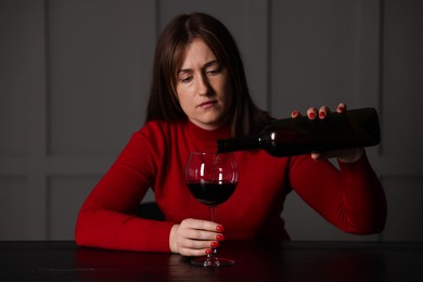 Photo of Alcohol addiction. Woman pouring red wine into glass at wooden table indoors