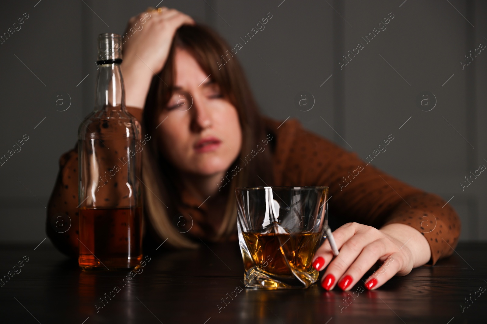 Photo of Alcohol addiction. Woman with glass of whiskey, cigarette and bottle at table indoors, selective focus