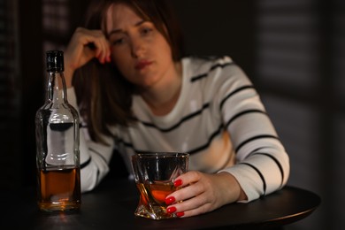 Photo of Alcohol addiction. Woman with glass of whiskey and bottle at table indoors, selective focus