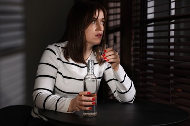 Photo of Alcohol addiction. Woman with shot glass of vodka and bottle at table indoors