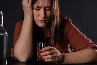 Photo of Alcohol addiction. Miserable woman with vodka at table in dark, closeup