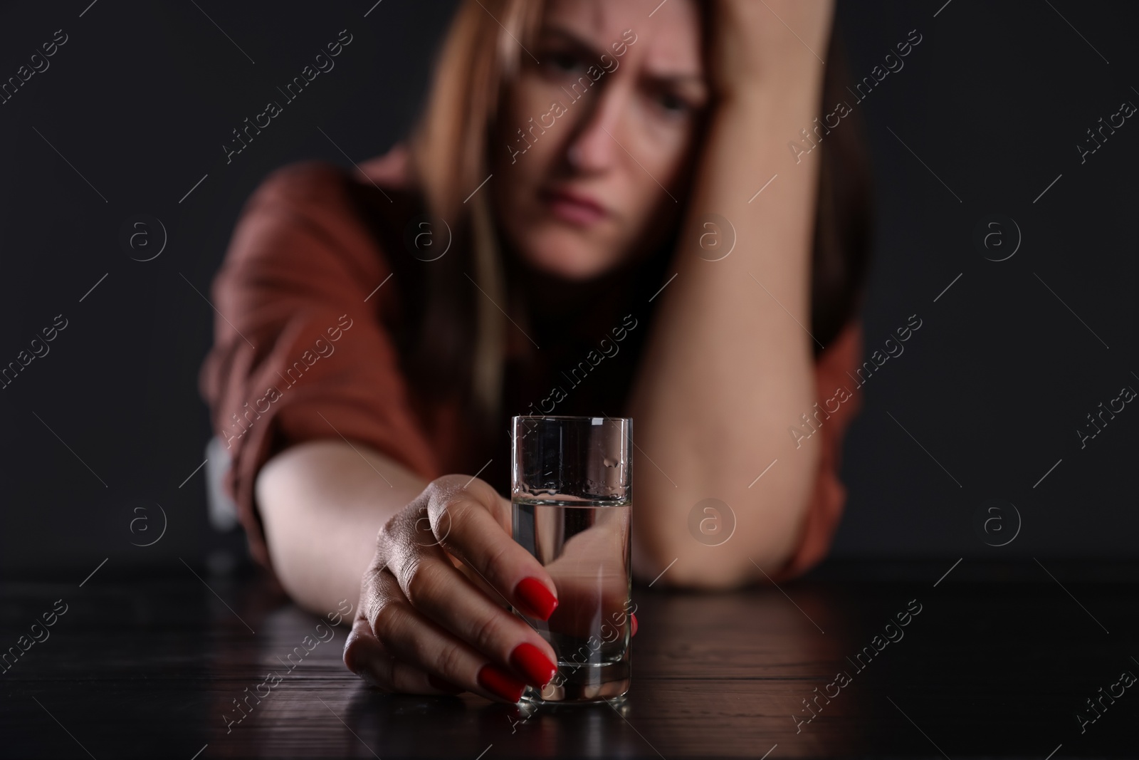 Photo of Alcohol addiction. Miserable woman with vodka at table in dark, selective focus