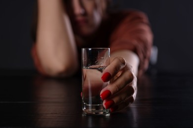 Photo of Alcohol addiction. Miserable woman with vodka at table in dark, closeup