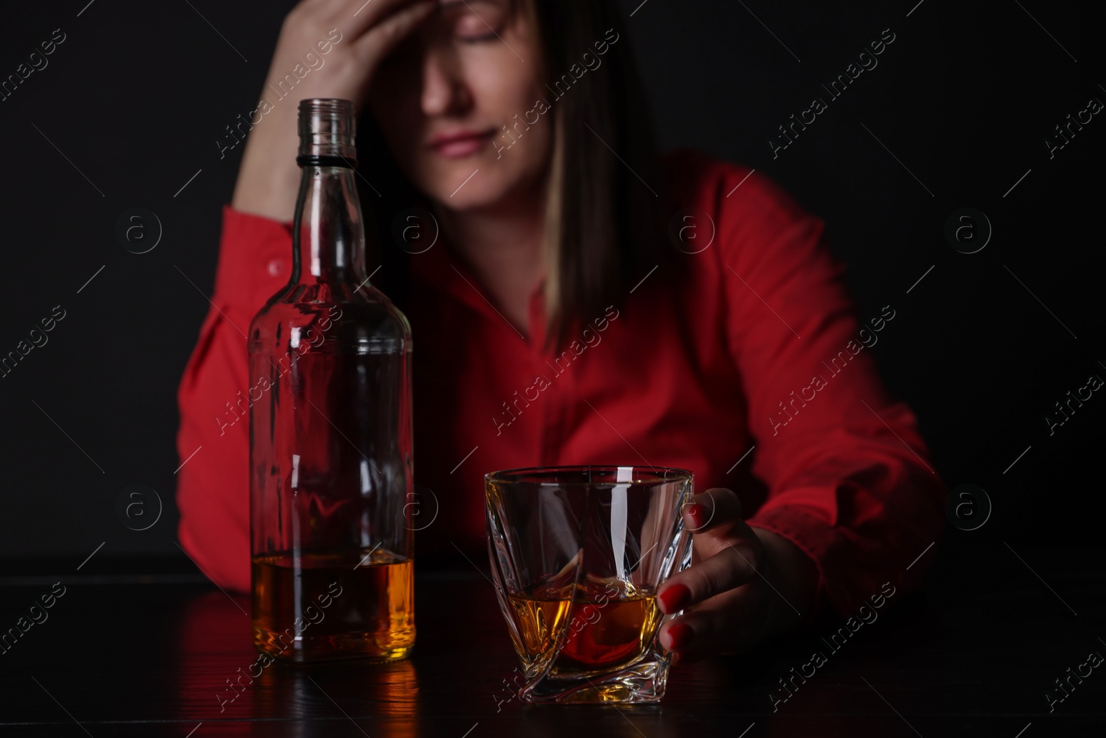 Photo of Alcohol addiction. Miserable woman with whiskey at table in dark, selective focus