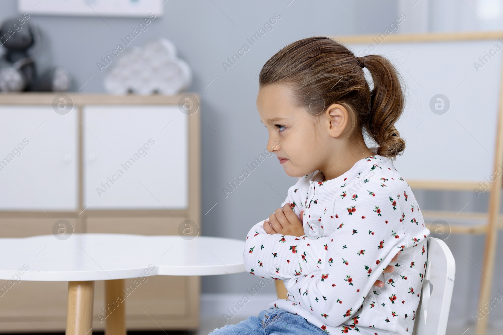 Photo of Resentful little girl at white table indoors, space for text