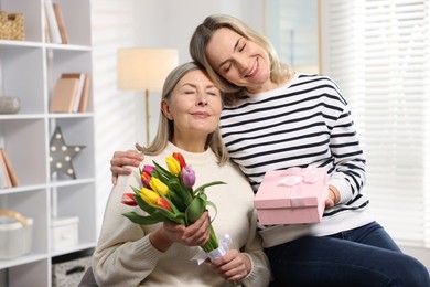 Photo of Smiling daughter congratulating her mom with bouquet of tulips and gift at home. Happy Mother's Day