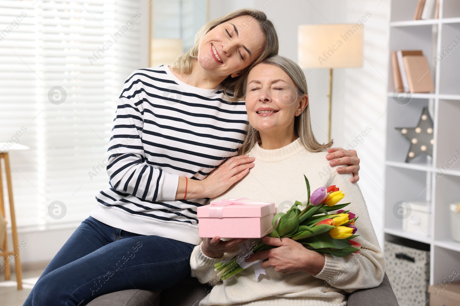 Photo of Smiling daughter congratulating her mom with bouquet of tulips and gift at home. Happy Mother's Day