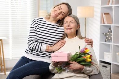 Photo of Smiling daughter congratulating her mom with bouquet of tulips and gift at home. Happy Mother's Day