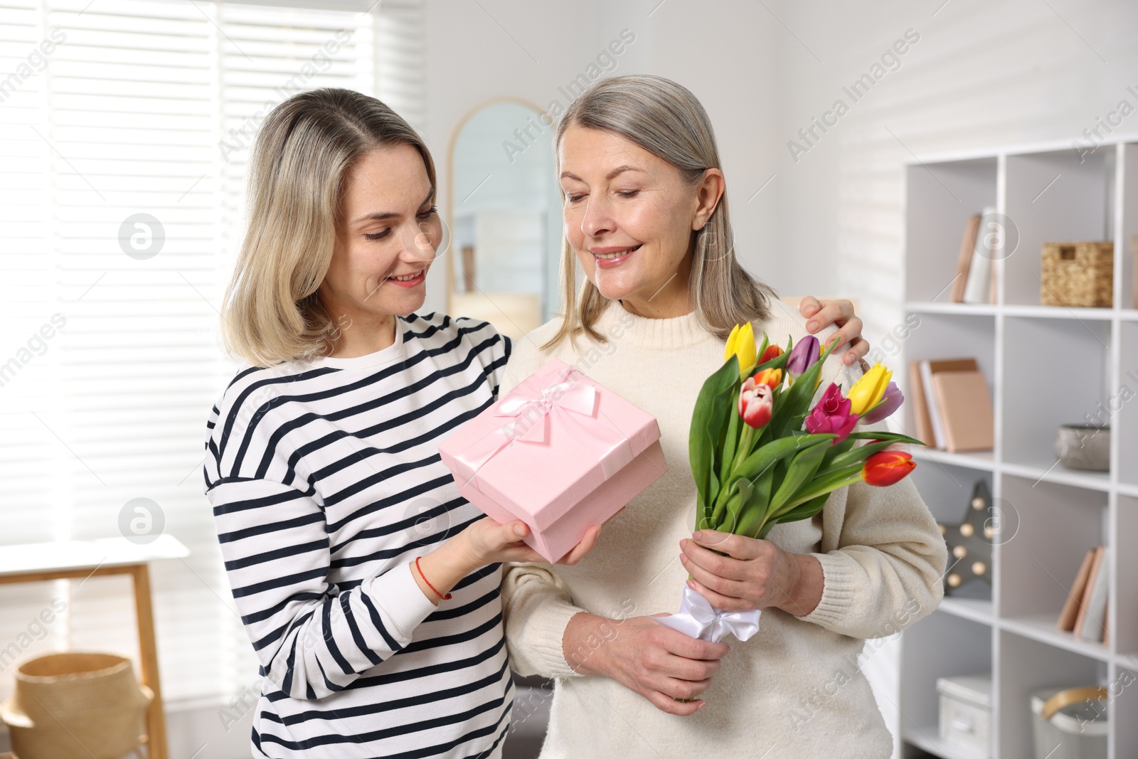 Photo of Smiling daughter congratulating her mom with bouquet of tulips and gift at home. Happy Mother's Day