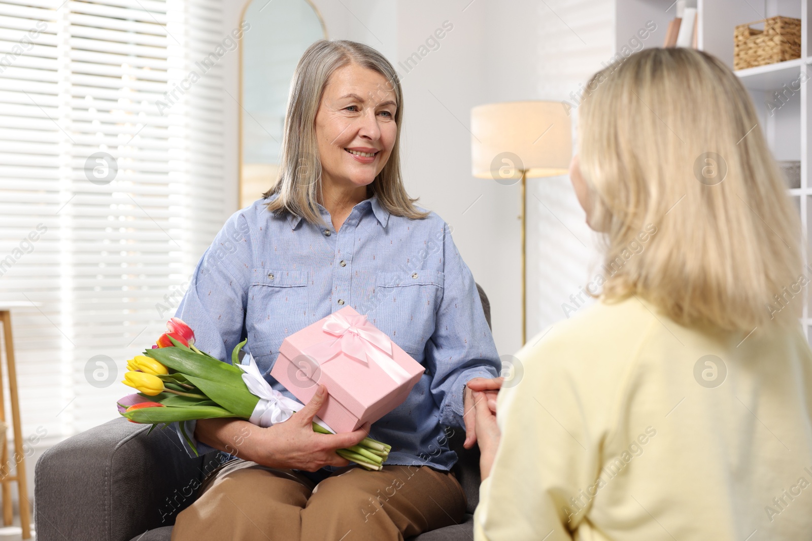 Photo of Smiling mom receiving bouquet of tulips and gift from her daughter on armchair at home. Happy Mother's Day