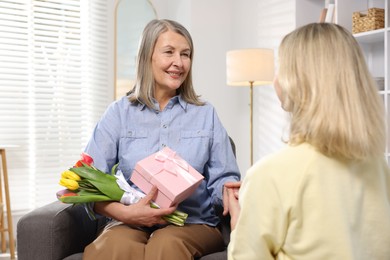 Smiling mom receiving bouquet of tulips and gift from her daughter on armchair at home. Happy Mother's Day