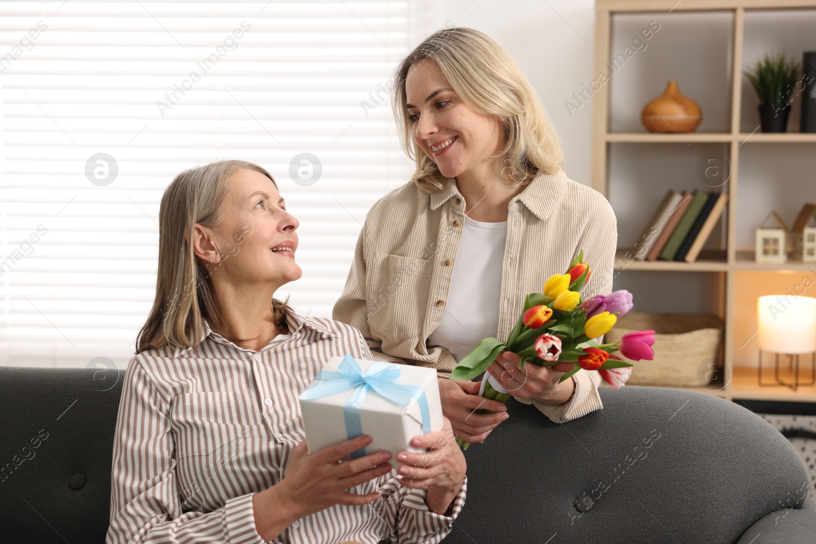 Photo of Smiling daughter congratulating her mom with bouquet of tulips and gift at home. Happy Mother's Day