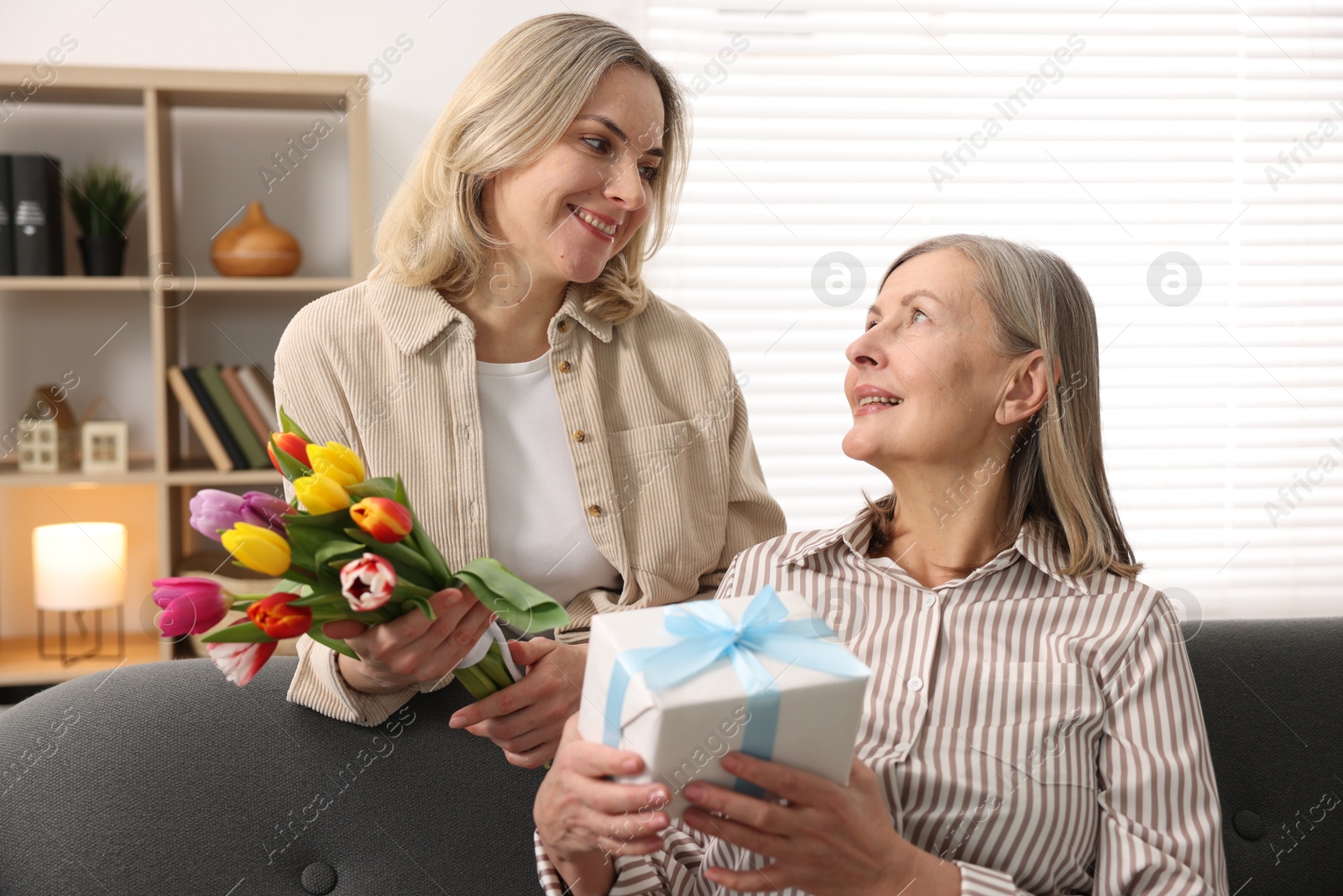 Photo of Smiling daughter congratulating her mom with bouquet of tulips and gift at home. Happy Mother's Day