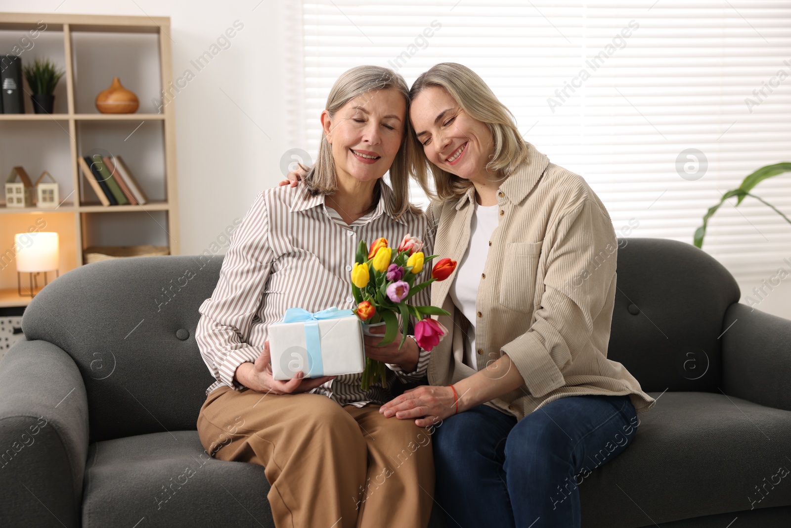 Photo of Smiling woman with bouquet of tulips, gift and her daughter on sofa at home. Happy Mother's Day