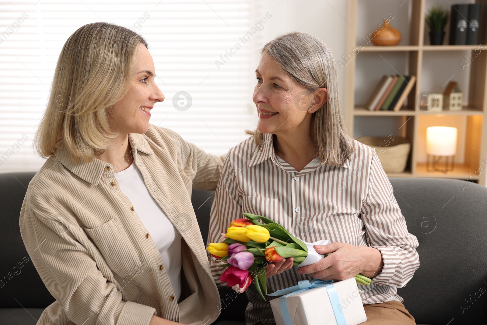 Photo of Smiling woman with bouquet of tulips, gift and her daughter on sofa at home. Happy Mother's Day