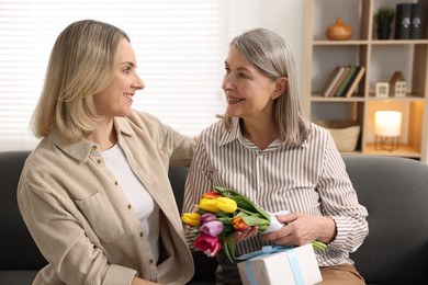 Photo of Smiling woman with bouquet of tulips, gift and her daughter on sofa at home. Happy Mother's Day