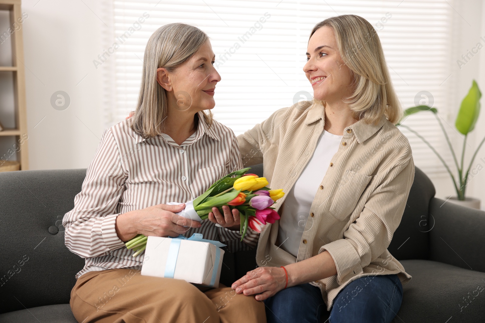 Photo of Woman with bouquet of tulips, gift and her smiling daughter on sofa at home. Happy Mother's Day