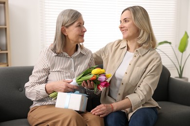 Woman with bouquet of tulips, gift and her smiling daughter on sofa at home. Happy Mother's Day