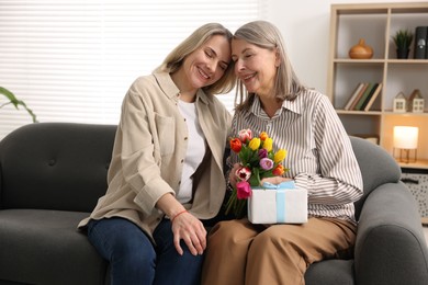 Photo of Smiling woman with bouquet of tulips, gift and her daughter on sofa at home. Happy Mother's Day