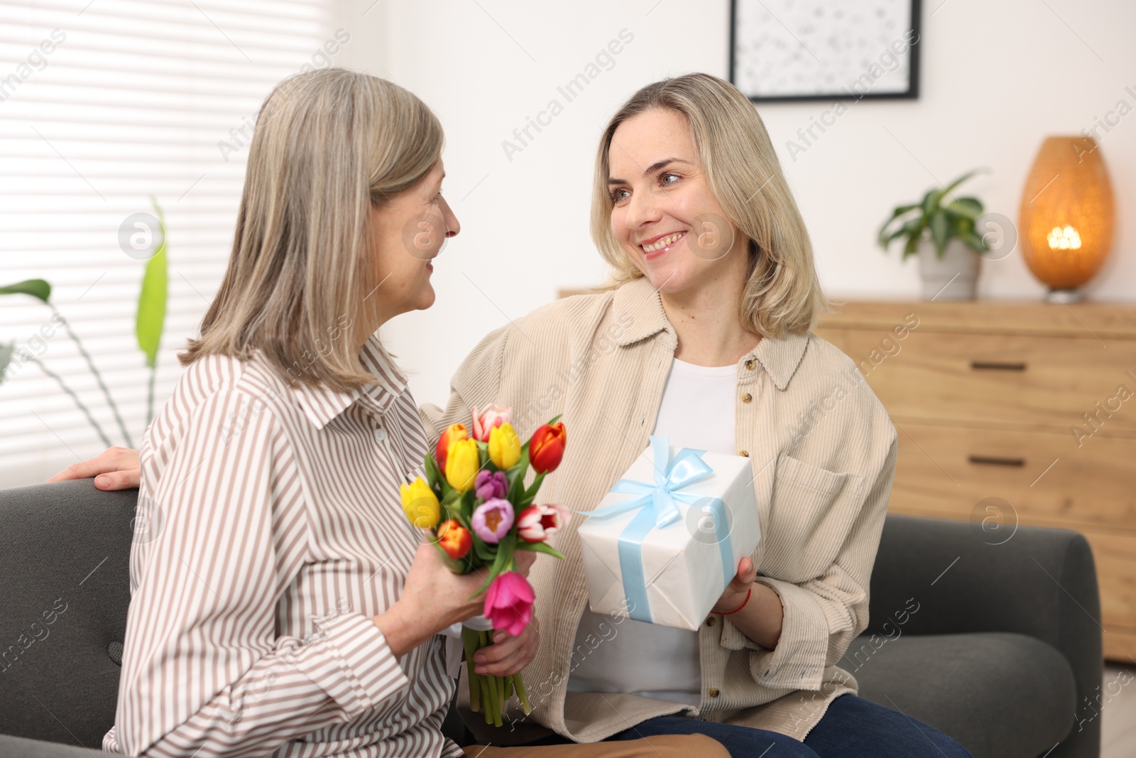 Photo of Smiling daughter congratulating her mom with bouquet of tulips and gift on sofa at home. Happy Mother's Day