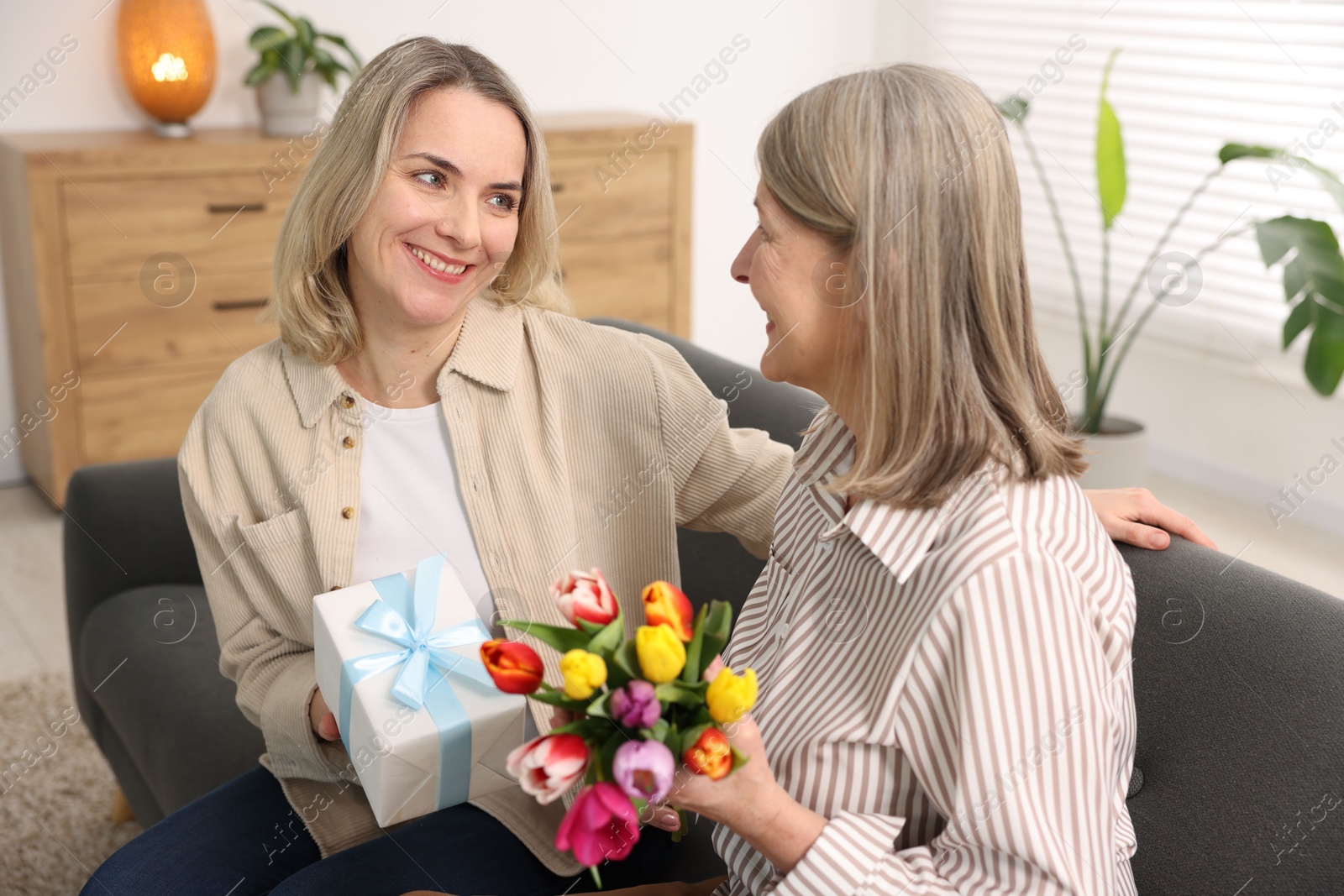 Photo of Smiling daughter congratulating her mom with bouquet of tulips and gift on sofa at home. Happy Mother's Day