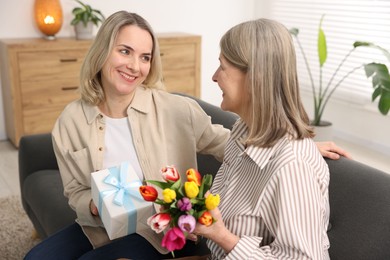 Photo of Smiling daughter congratulating her mom with bouquet of tulips and gift on sofa at home. Happy Mother's Day