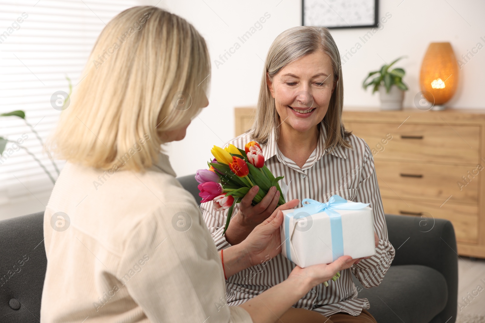 Photo of Smiling mom receiving bouquet of tulips and gift from her daughter on sofa at home. Happy Mother's Day