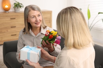 Smiling mom receiving bouquet of tulips and gift from her daughter on sofa at home. Happy Mother's Day