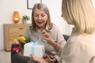 Photo of Smiling mom receiving bouquet of tulips and gift from her daughter on sofa at home. Happy Mother's Day