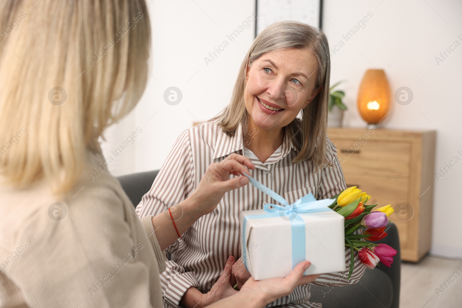 Photo of Smiling mom receiving bouquet of tulips and gift from her daughter on sofa at home. Happy Mother's Day