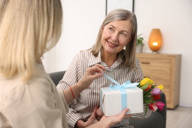 Photo of Smiling mom receiving bouquet of tulips and gift from her daughter on sofa at home. Happy Mother's Day