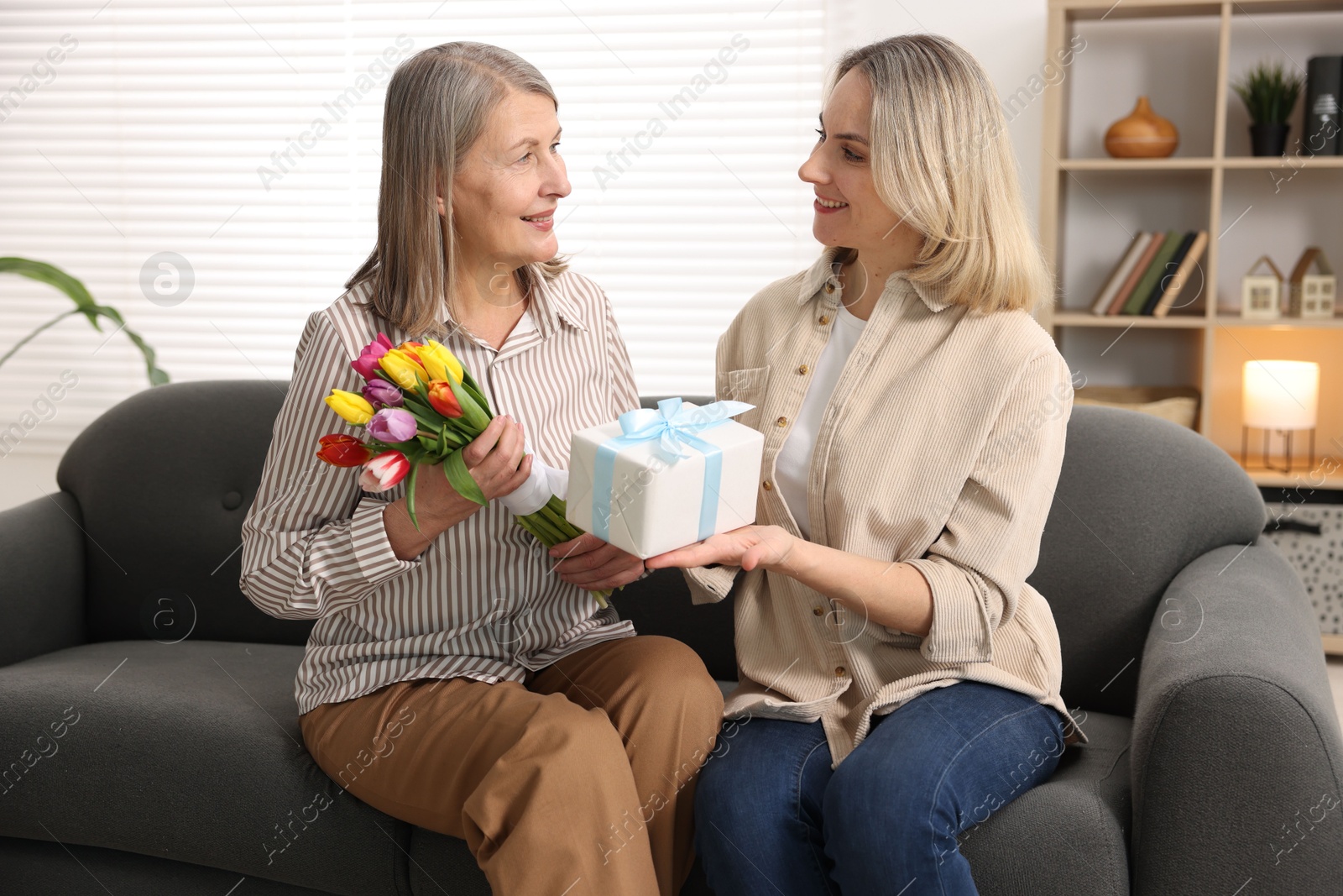 Photo of Smiling daughter congratulating her mom with bouquet of tulips and gift on sofa at home. Happy Mother's Day