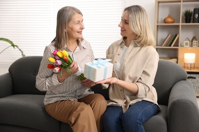 Photo of Smiling daughter congratulating her mom with bouquet of tulips and gift on sofa at home. Happy Mother's Day